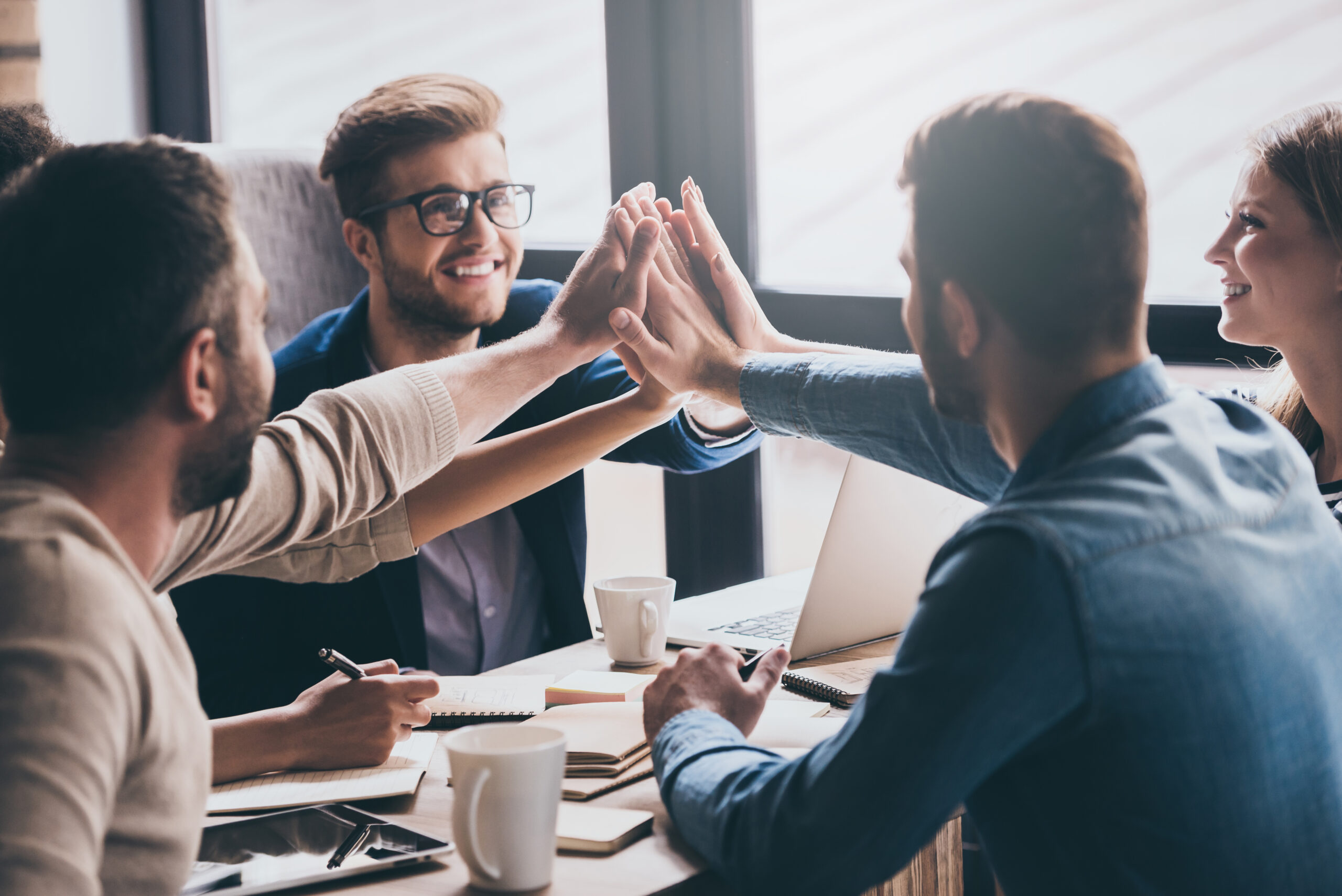 We are leaders on the market! Cheerful young people giving each other high-five with smile while sitting at the office table on business meeting